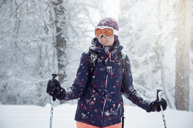 Giovane sciatrice in piedi sulla pista da sci sotto la nevicata che tiene i bastoncini da sci e sorride da vicino