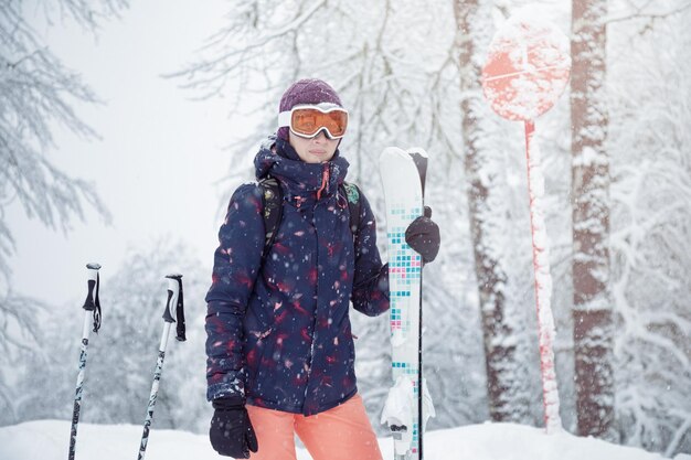 Giovane sciatrice in piedi sulla pista da sci sotto la nevicata che tiene i bastoncini da sci e sorride da vicino