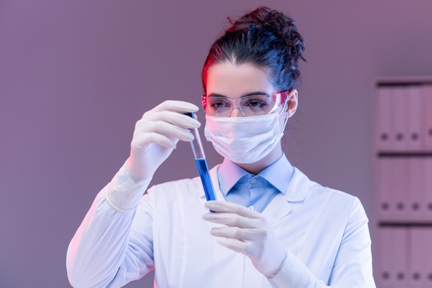 Giovane scienziato femminile in maschera, guanti e occhiali da vista guardando il pallone mentre studiava la sostanza liquida blu in laboratorio