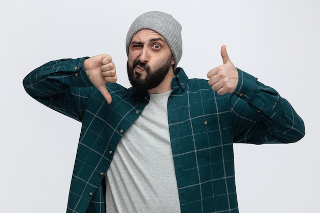 Giovane scontento che indossa un cappello invernale guardando la fotocamera che mostra i pollici su e giù isolati su sfondo bianco