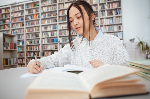 Giovane scrittura asiatica della studentessa in suo taccuino alla biblioteca, libro aperto su priorità alta