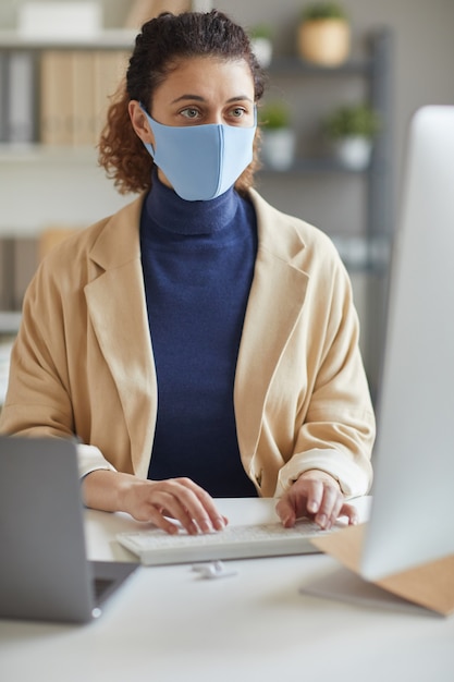 Giovane segretario in maschera protettiva seduto al suo posto di lavoro digitando sulla tastiera del computer che lavora in ufficio