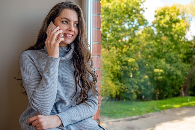 Giovane sensuale bruna dai capelli lunghi millenari con lo smartphone seduto sul davanzale della finestra a casa