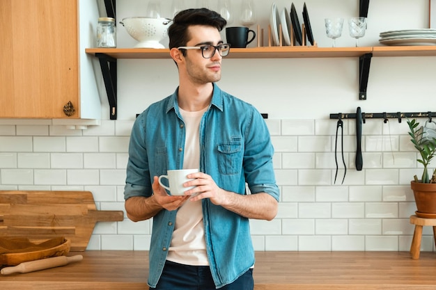 Giovane serio che beve tè o caffè in cucina preoccupato per la situazione nel mondo