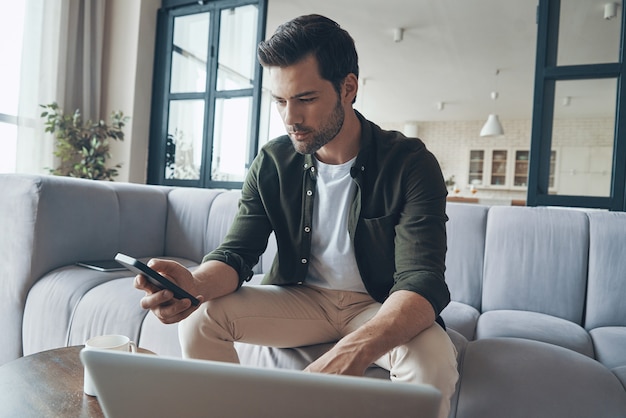 Giovane serio che utilizza smartphone e laptop mentre è seduto sul divano