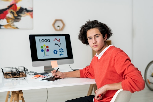 Giovane serio web designer con stilo e tavoletta grafica seduto alla scrivania davanti alla telecamera in ambiente domestico
