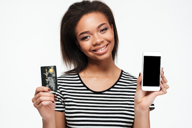 Giovane signora africana felice che per mezzo del telefono e tenendo la carta di debito.