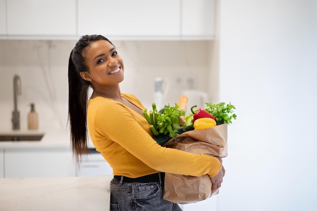 Giovane signora afroamericana allegra che tiene sacchetto di carta con generi alimentari nello spazio libero interno della cucina moderna