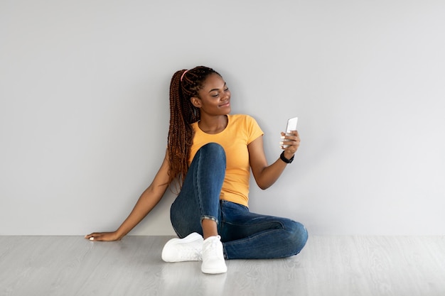 Giovane signora afroamericana seduta con il cellulare che naviga in internet in chat online contro il grigio