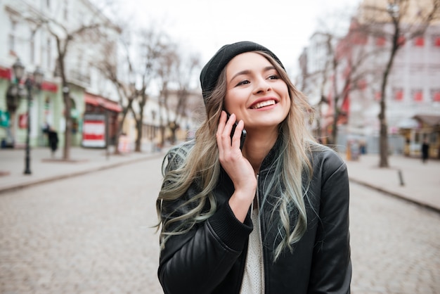 Giovane signora allegra che cammina sulla via e che parla dal telefono.