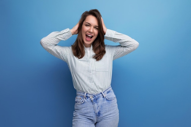 Giovane signora castana ben curata vestita con una camicia a righe e jeans sorridenti su uno sfondo di studio