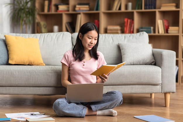 Giovane signora coreana che studia online da casa leggendo un libro e usando il pc portatile seduto sul pavimento vicino