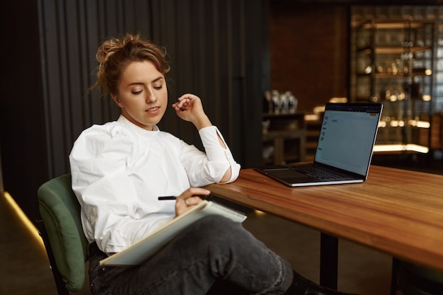 Giovane signora vaga in camicetta bianca e jeans neri che scrive le note mentre sedendosi al tavolo con il computer portatile aperto