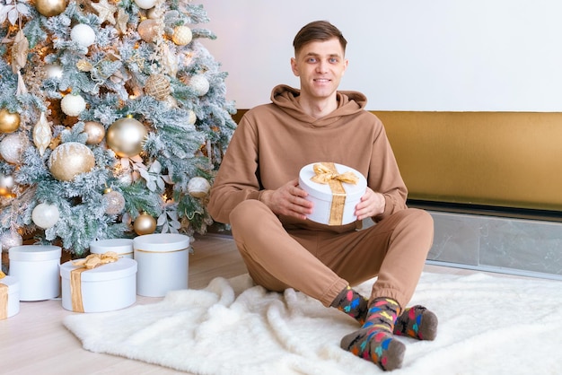 Giovane sorridente a Natale vicino all'albero di Natale seduto sul pavimento in un ambiente accogliente