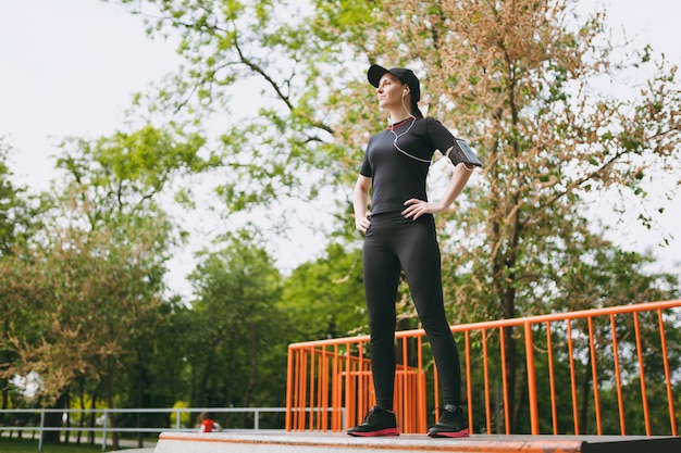 Giovane sorridente atletica bella donna bruna in uniforme nera, berretto con cuffie facendo esercizi sportivi, riscaldamento prima di correre, in piedi nel parco cittadino all'aperto