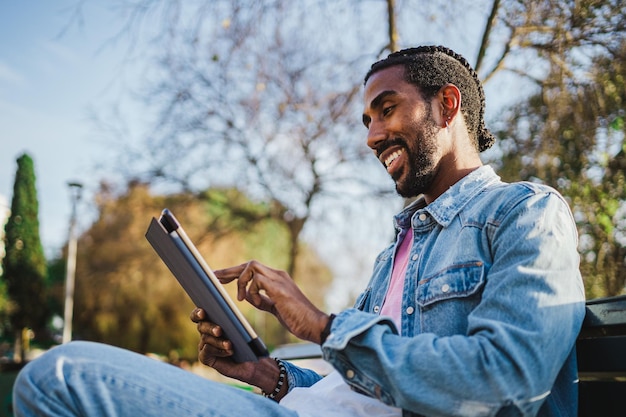 Giovane sorridente che legge un libro sulla sua tavoletta digitale È seduto su una panchina nel parco