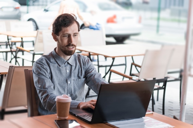 Giovane sorridente che si siede al tavolo nella caffetteria che lavora al computer portatile.