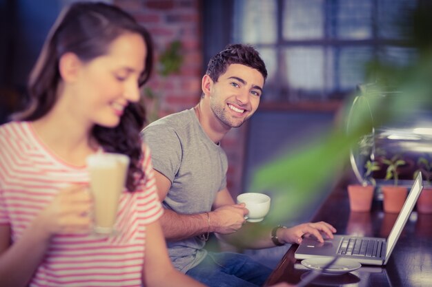 Giovane sorridente che si siede alla barra e che per mezzo del computer portatile