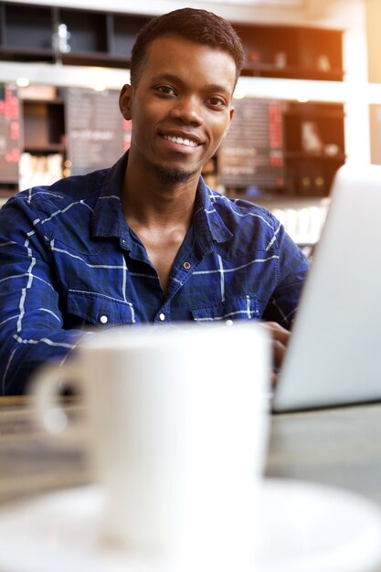 Giovane sorridente che si siede in caffè con la tazza di caffè e il computer portatile