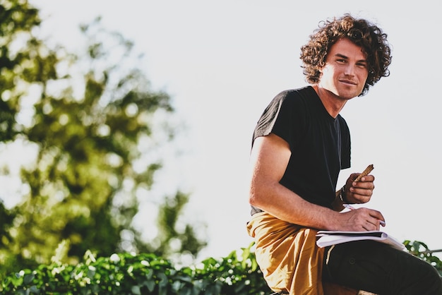 Giovane sorridente con i capelli ricci che scrive alcuni avvisi su carta e si prepara per gli esami indossando una maglietta nera seduto sulla strada della città L'uomo d'affari freelance fa piani per nuovi progetti