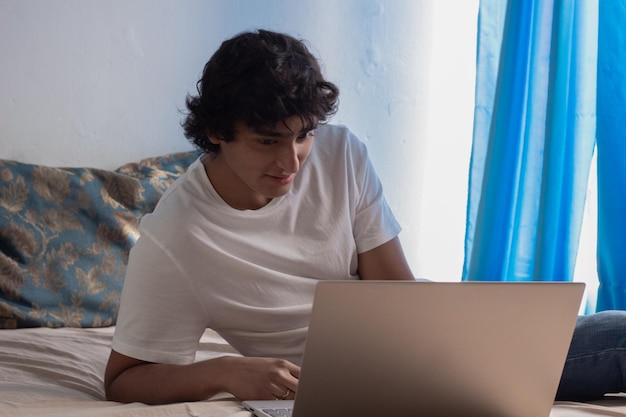 Giovane sorridente con i capelli ricci sdraiato sul letto che lavora al suo computer portatile da casa