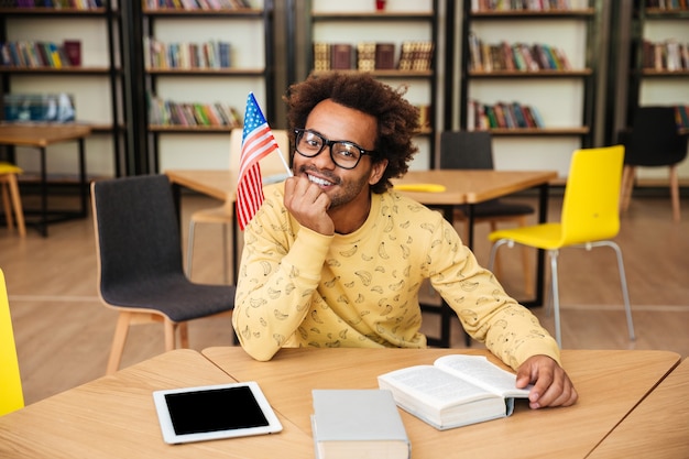 Giovane sorridente con la bandiera che si siede e che legge il libro in biblioteca