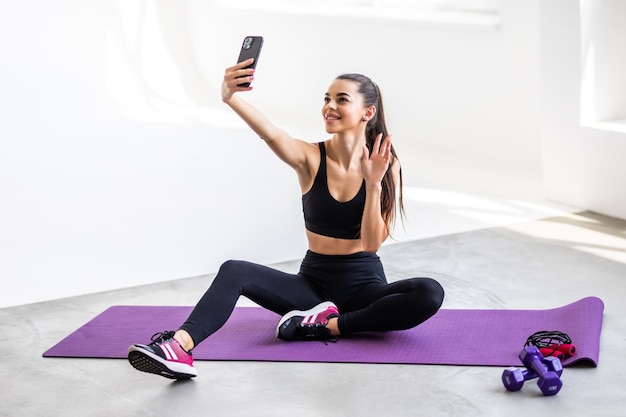 Giovane sorridente forte sportivo atletico fitness trainer istruttore donna facendo esercizi fare selfie girato sul cellulare si siede sul pavimento della stuoia a casa palestra