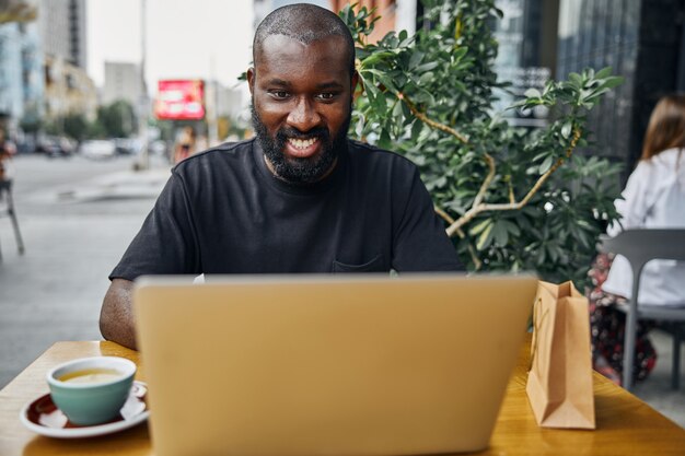 Giovane sorridente positivo che guarda lo schermo di un laptop moderno mentre è seduto su una terrazza di un caffè all'aperto