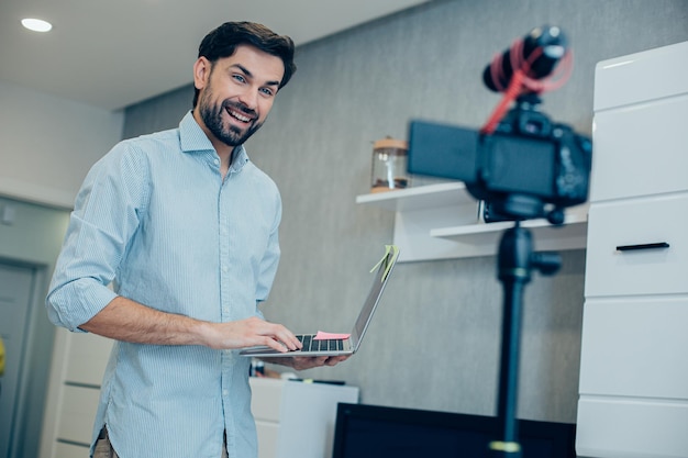 Giovane specialista entusiasta in piedi con un laptop moderno davanti a una telecamera e sorridente durante la registrazione di un video