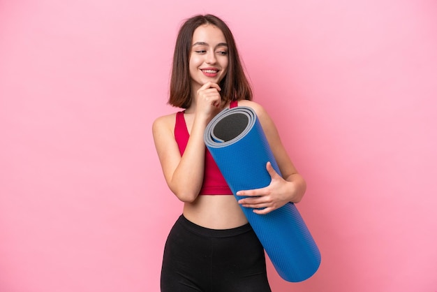 Giovane sport Donna ucraina che va a lezioni di yoga mentre tiene un tappetino isolato su sfondo rosa guardando di lato e sorridente