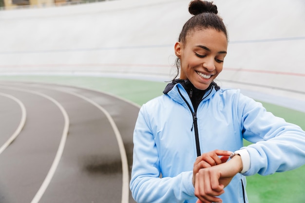 giovane sportiva africana che riposa dopo aver corso allo stadio, guardando smartwatch