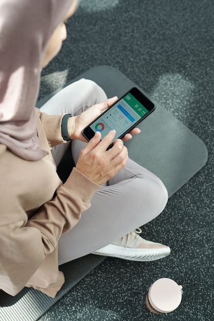 Giovane sportiva in hijab e abbigliamento sportivo che guarda attraverso i suoi progressi di fitness nel telefono cellulare