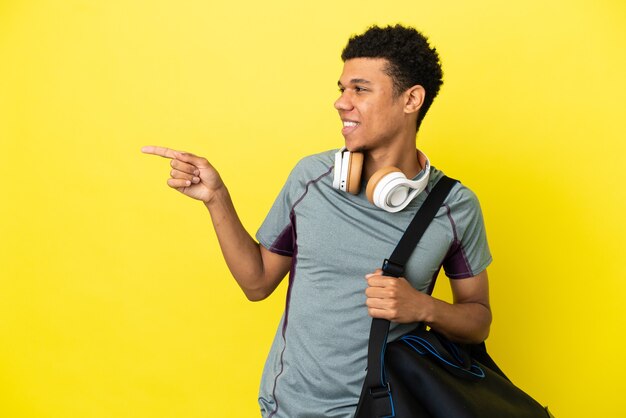 Giovane sportivo afroamericano con borsa sportiva isolata su sfondo giallo che punta il dito di lato e presenta un prodotto