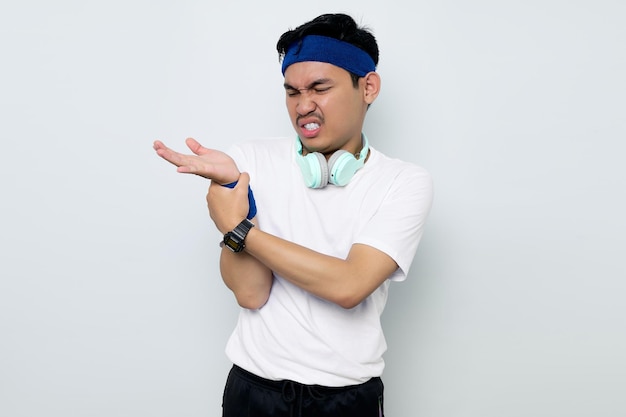 Giovane sportivo asiatico forte in fascia blu e maglietta bianca da abbigliamento sportivo con le cuffie che tengono il braccio del polso soffre di dolore isolato su sfondo bianco Concetto di sport di allenamento