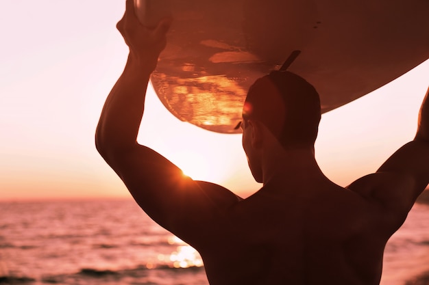 Giovane sportivo con tavola da surf sulla spiaggia al tramonto