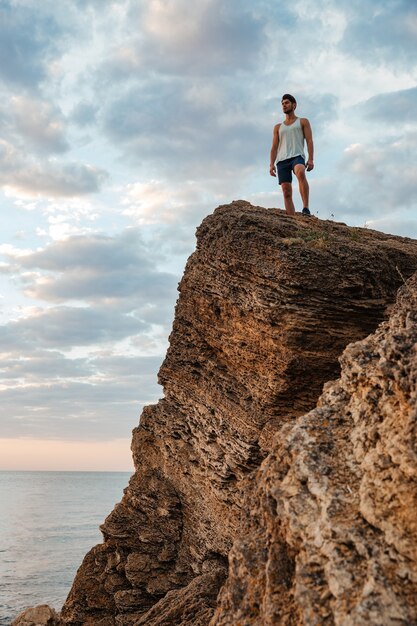 Giovane sportivo in piedi sulla roccia di montagna in riva al mare e guardando il tramonto