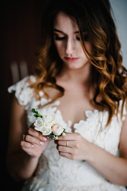 Giovane sposa attraente che tiene l'asola dello sposo. fiori con rose rosa, garofani bianchi e verdi e verdi. I preparativi della sposa. Mattina di nozze.