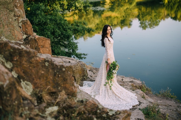 Giovane sposa in piedi sulla riva rocciosa del fiume, tramonto