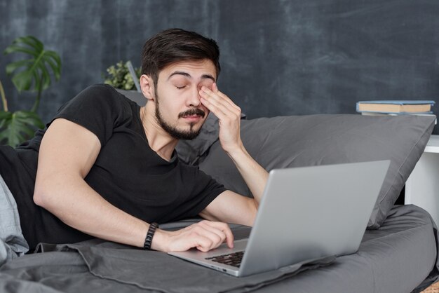 Giovane stanco che ha affaticamento degli occhi sdraiato sul letto con il computer portatile e lo sfregamento degli occhi