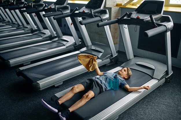 Giovane stanco sdraiato sul tapis roulant in palestra, macchina da corsa. Ragazzo in formazione, assistenza sanitaria e stile di vita sano, scolaro in allenamento