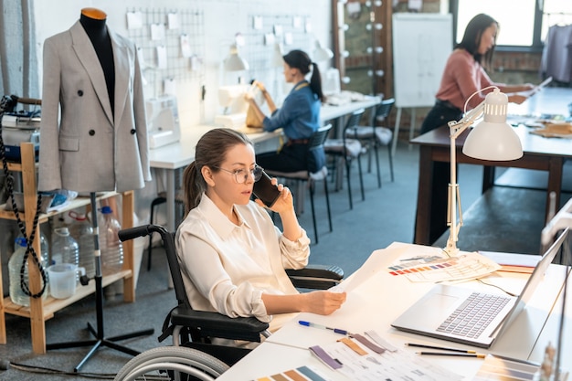 Giovane stilista femminile che parla al telefono cellulare durante il lavoro davanti al laptop