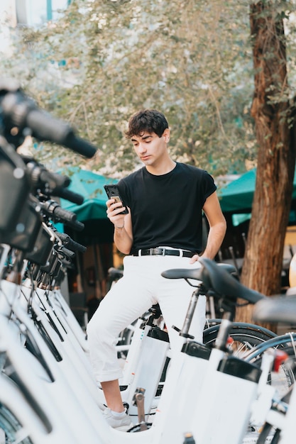 Giovane studente adulto che noleggia una bicicletta con il suo smartphone. Vestiti moderni usando il suo telefono vicino a un servizio di noleggio biciclette, sbloccando biciclette a noleggio per spostarsi in città, servizi di noleggio, spazio di copia