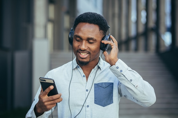 Giovane studente africano che ascolta musica dallo smartphone usando grandi cuffie, sorridendo per la comodità di usare l'app