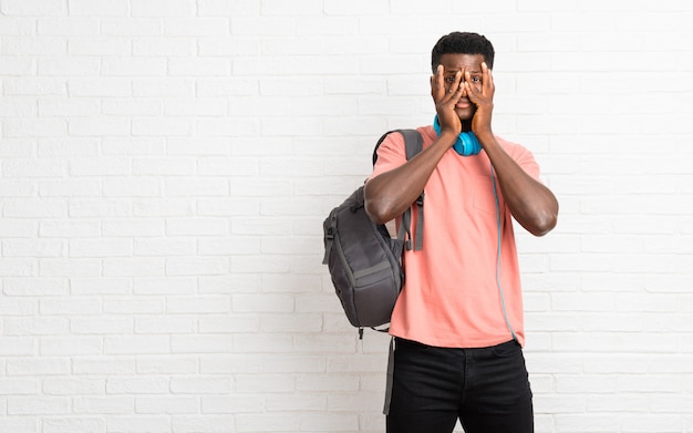 Giovane studente afro americano uomo sorpreso e che copre il viso con le mani mentre guardando attraverso le dita