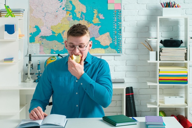 Giovane studente alla moda con i libri sullo sfondo dell'aula in chirt blu che mangia il preparatore dello studente della mela