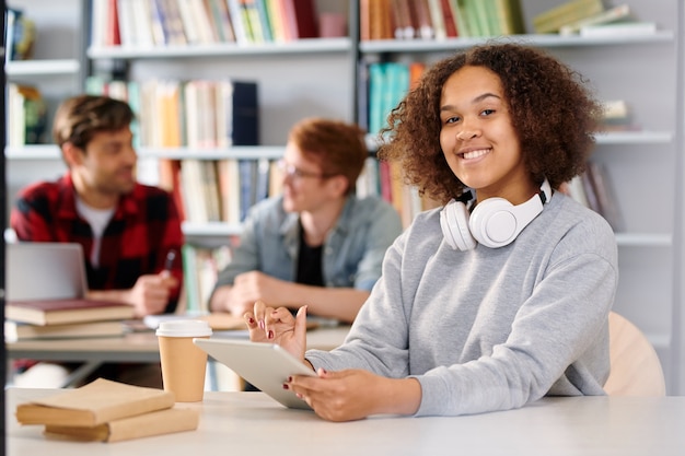 Giovane studente allegro con tablet si prepara per l'esame mentre si lavora nella biblioteca del college