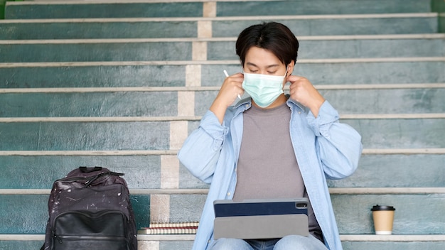 Giovane studente asiatico del campus che indossa una maschera di protezione durante lo studio online nel campus, prevenzione del coronavirus all'università, distanza sociale, apprendimento online