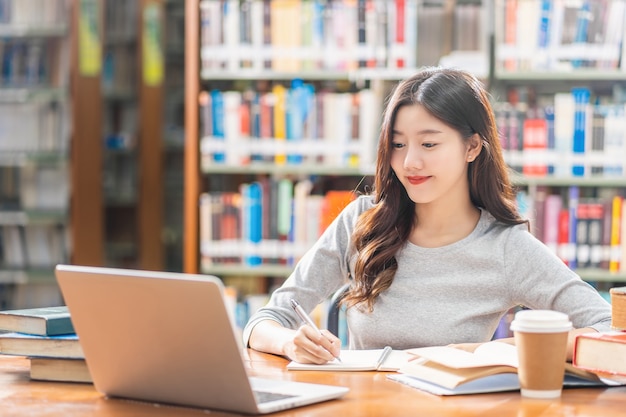 Giovane studente asiatico in abito casual che fa i compiti e usa il computer portatile tecnologico in biblioteca