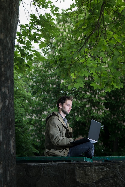 Giovane studente bello che si siede su una panchina di legno nel parco con il computer portatile.