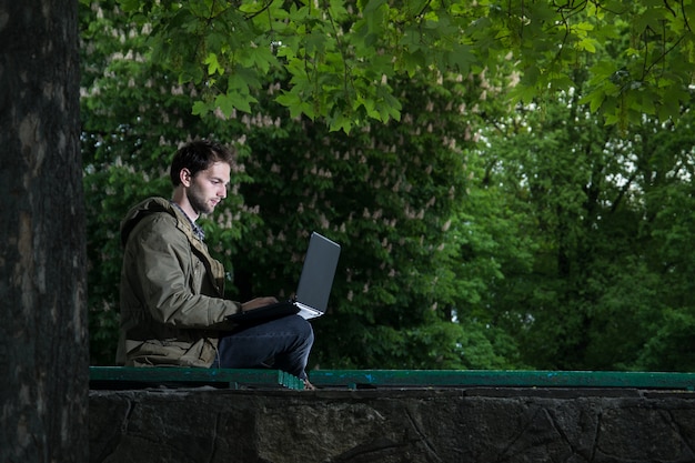 Giovane studente bello che si siede su una panchina di legno nel parco con il computer portatile.